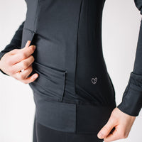 Woman showing kangaroo pocket of black maternity and nursing hoodie with hidden zippers, large hood, thumb holes and hand covers.