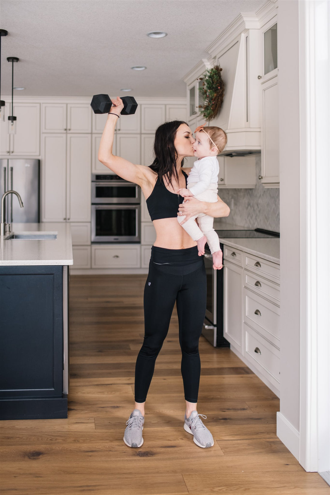 Mother holding baby while working out in black front-zip nursing sports bra and black maternity leggings.