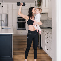 Mother holding baby while working out in black front-zip nursing sports bra and black maternity leggings.
