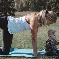 Mother doing yoga with baby, wearing lavender maternity tank top and black maternity leggings from Joyleta.